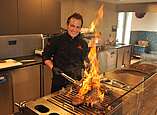 Ein Koch grillt Fleisch auf einem Grill, aus diesem kommt eine große Flamme.
