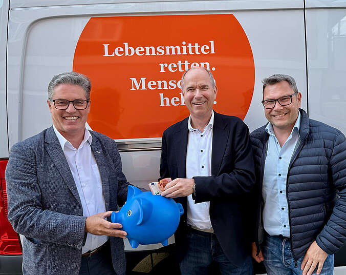 Drei Männer mit blauem Sparschwein vor Tafel-Transportwagen