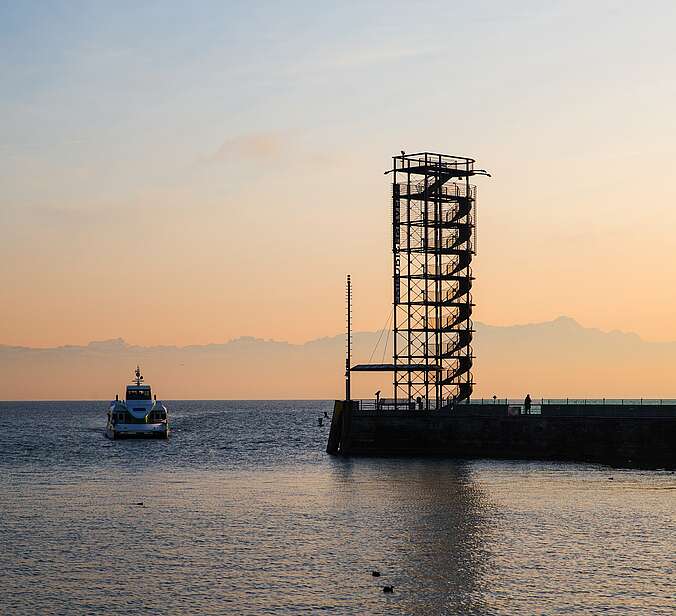 Hafeneinfahrt im Sonnenuntergang mit Blick auf den Moleturm in Friedrichshafen und ein einfahrendes Schiff.