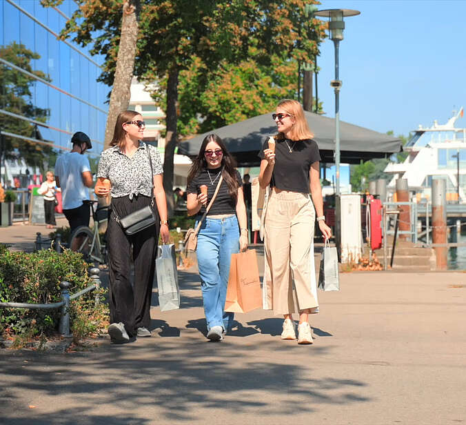 Screenshot aus Film: Drei junge Frauen beim Shoppen mit Tüten. Sie schlendern entlang der Uferpromenade.