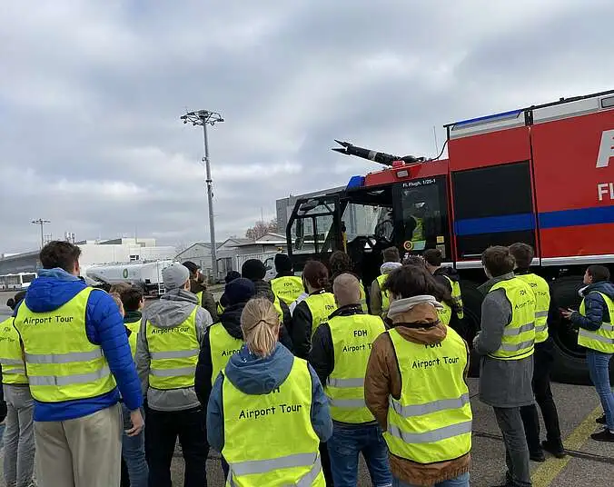 Foto mit Menschen in Warnwesten neben einem Flughafen Feuerwehrauto