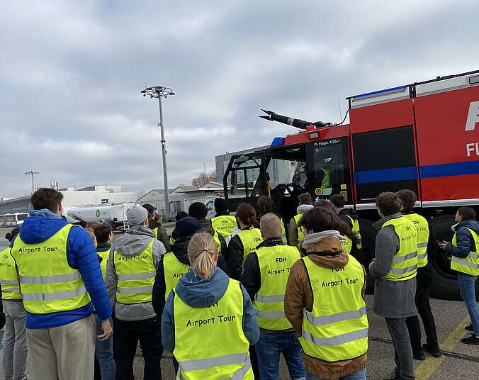 Foto mit Menschen in Warnwesten neben einem Flughafen Feuerwehrauto