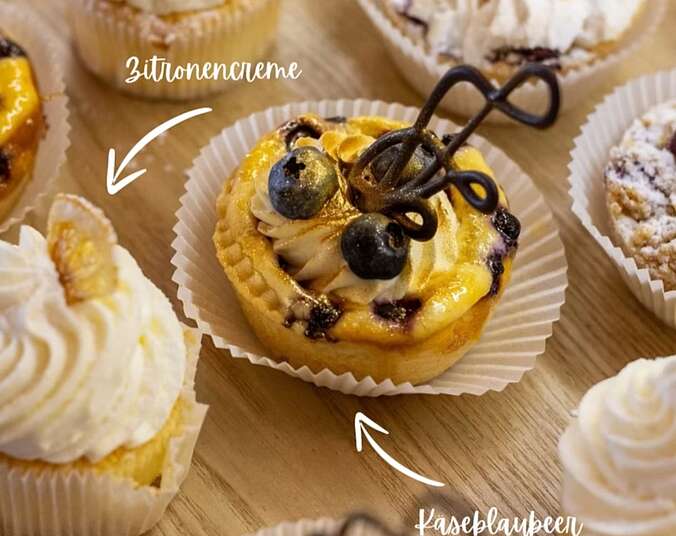 Nahaufnahme verschiedener kleiner Tartelettes auf einer Fläche nebeneinander. Verziert mit aufdressierten Sahnehäubchen, Blaubeeren und kandierten Zitronenstückchen.