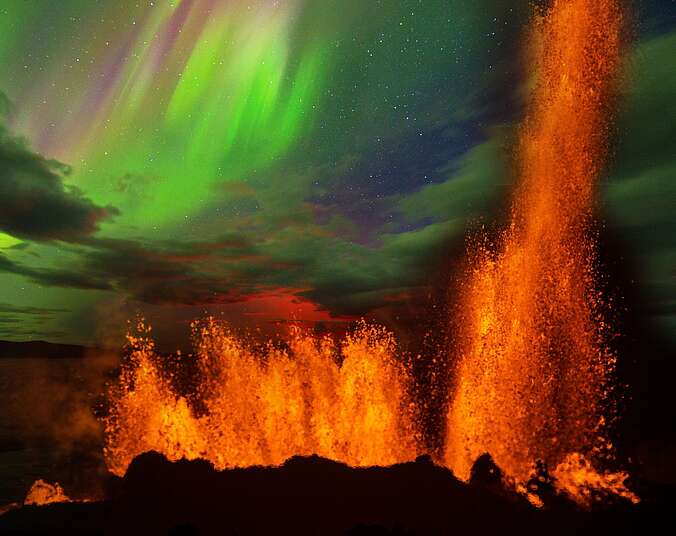 Eine orange glühende Lavafontäne spritzt auf dem Boden, am Himmel im Hintergrund sind grün leuchtende Polarlichter zu sehen.