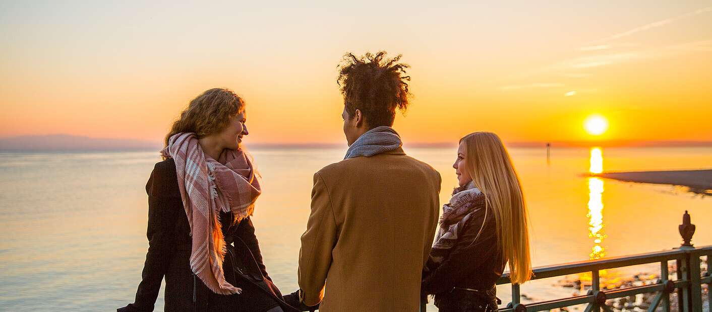 Drei Menschen schauen am See zum Sonnenuntergang