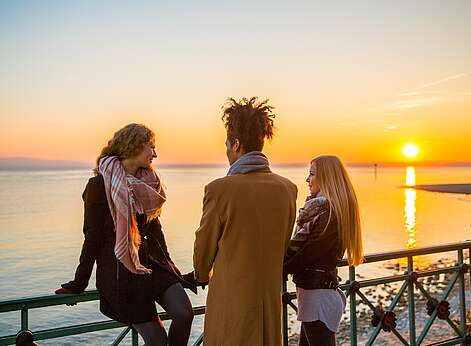 Drei Menschen schauen am See zum Sonnenuntergang