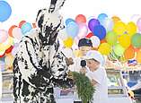 Mann in Hasenkostüm übernimmt von zwei Kindern ein Bund Karotten im Hintergrund Luftballons