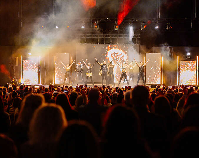 Bild aus dem Zuschauerraum auf eine Bühne mit projeziertem Bühnenbild und Pyrotechnik und ungefähr neun Tänzerinnen und Tänzern in V-Formation.