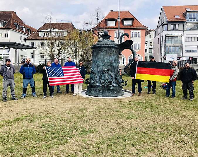 Übergroßer Krug aus Bronze steht auf Wiese, Menschen rechts und links daneben mit Deutschland-Flagge und Flagge der USA