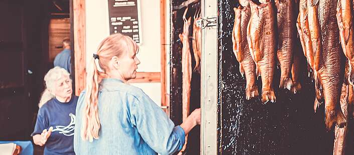 Eine Frau steht vor einem offenen Räucherofen. In diesem hängen geräucherte Fische.
