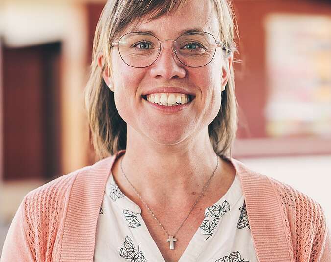 Eine junge Frau mit offenem Lächeln, Brille mit dezentem Gestell und schulterlangen blonden Haaren. Sie trägt eine weiße Bluse mit Schnetterlingen in Gittermuster und darüber offen eine rosafarbene Strickjacke.