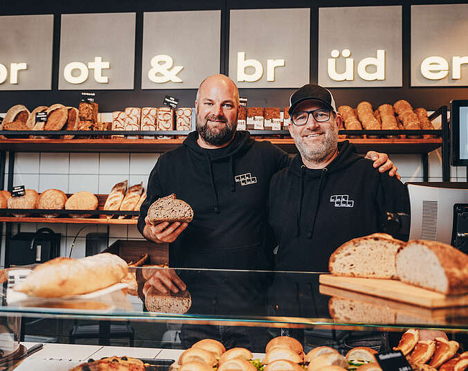 Zwei Männer stehen hinter einer Theke mit lecker angerichtetem Brot und Brötchen.
