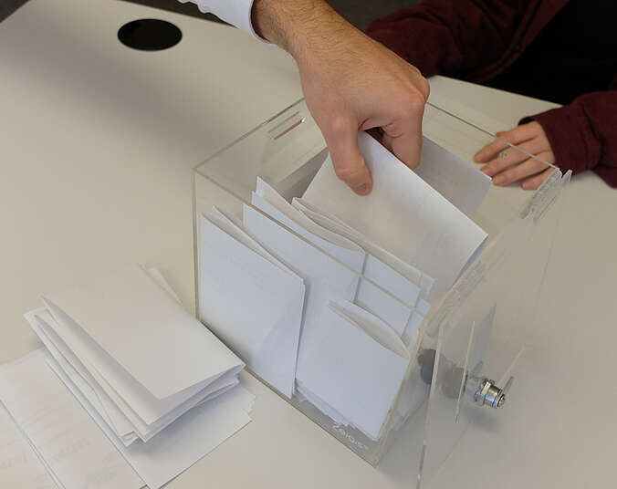 Hand greift in eine durchsichtige Box mit weißen Zetteln