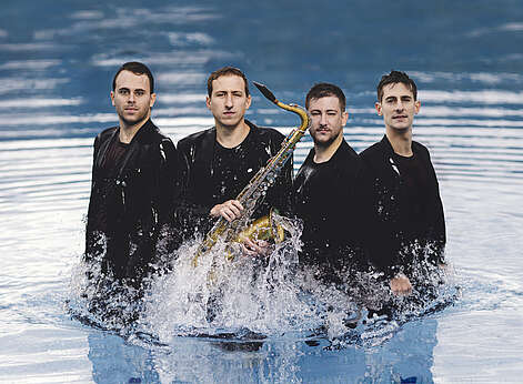 4 Männer stehen im Wasser - einer hält ein Saxophon.