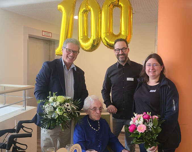 Männer und Frauen mit Blumen und einem Luftballon der eine 100 darstellt.