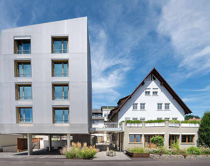Ein kleines weißes Haus neben einem modernen Neubau, mit Blumen und Büschen an der Vorderseite.
