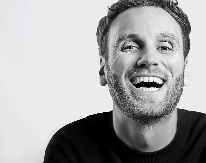 Schwarzweiß-Porträt eines Mannes mittleren Alters mit kurzen Haaren. Er lacht mit offem Mund.