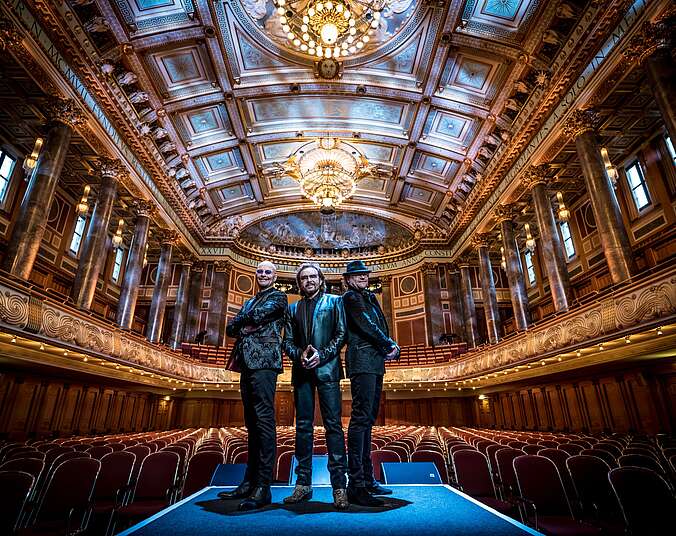 Drei Männer stehen komplett in Schwarz gekleidet auf einer Bühne mit dem Rücken zum Zuschauerraum eines prunkvollen Konzertsaals mit Samt bezogenen Stühlen, umlaufenenden Emporenrängen mit goldenen Säulen und Kornleuchtern.