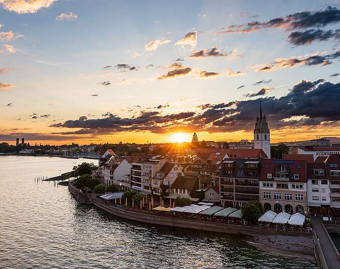 Blick vom See auf die Uferpromenade und die Stadt Friedrichshafen mit der Nikolauskirche und im Hintergrund die Schlosskirche bei Sonnenuntergang.