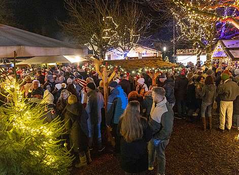 Frauen und Maener stehen im weihnachtlich beleuchteten Winterdorf im Lammgarten an runden Stehtischen.