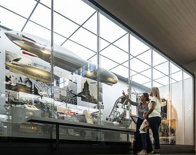 In der Fotografie ist eine Familie zu sehen, die im Zeppelin Museum vor einer großen Vitrine steht und unterschiedliche Zeppelin-Modelle betrachtet.