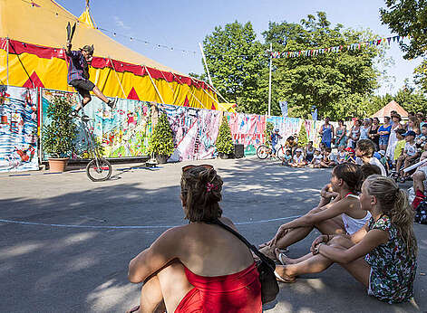 Zuschauer sitzen auf der Straße und bestaunen einen Akrobaten auf einem hohen Einrad.
