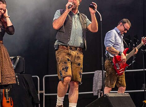 Zwei Männer in Lederhosen stehen auf einer Bühne - einer mit Mikrofon, der andere mit E-Gitarre.
