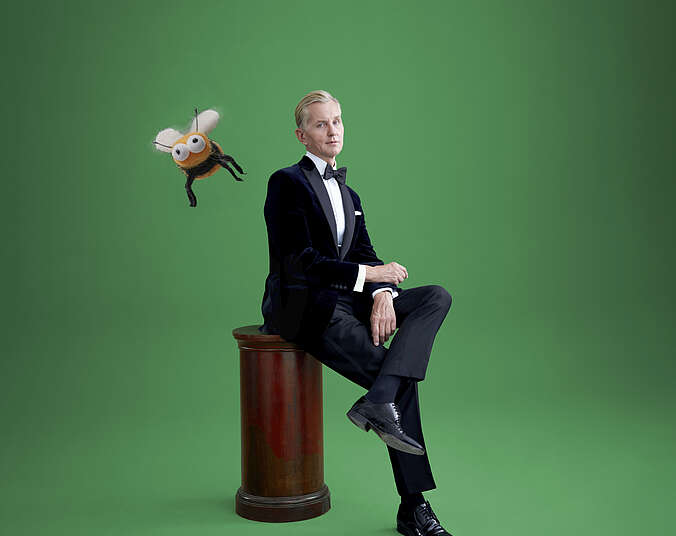 Ein Mann mit blonden, zurückgegelten Haaren in schwarzen Smoking mit Fliege sitzt vor einem grünen Hintergrund mit übereinandergesclagenen Beinen auf einer dunkelbraunen Holzsäule. Links von ihm fliegt eine gezeichnete Hummel durch das Bild.