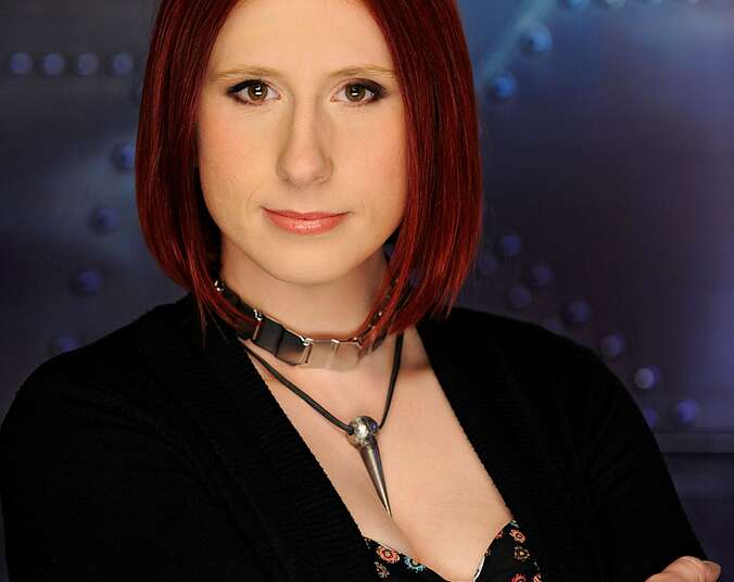 Eine junge Frau mit Pagenschnitt, mit verschränkten Armen, trägt ein dunkles Oberteil und zwei silberne massive Halsketten und blickt freundlich und frontal in die Kamera.