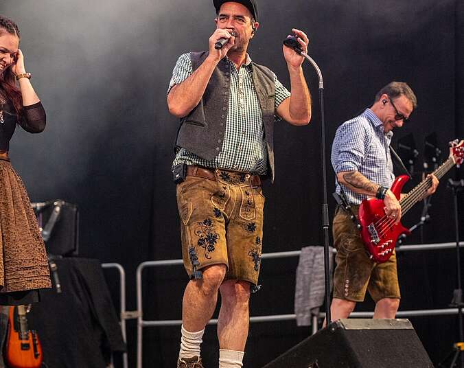 Zwei Männer in Lederhose stehen auf einer Bühne - einer mit Mikrofon in der Hand, der andere mit einer E-Gitarre.