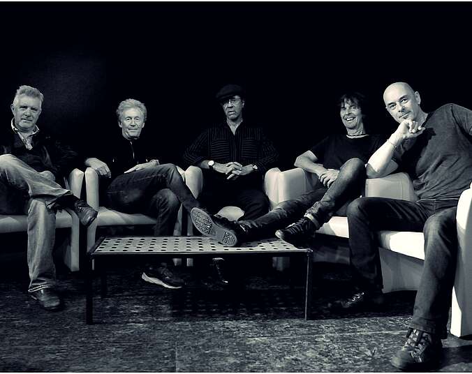 Schwarz-Weiß-Bild, auf dem fünf Männer in schwarzen T-Shirts und dunklen Jeanshosen in einem Halbkreis in fünf weißen Ledersesseln sitzen.