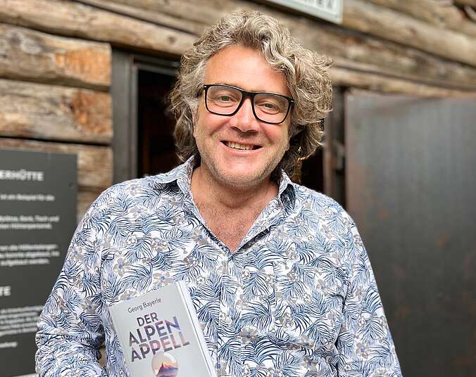 Georg Bayerle vor DAV Hütte mit Buch