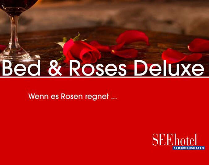 Der Stiel eines Weinglases mit Rotwein und eine rote Rose sowie Rosenblätter, die auf einem Tisch liegen.