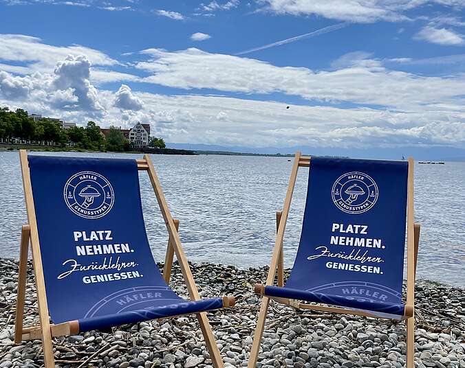 Zwei Lieestühle mit Aufschrift "Häfler Genusstypen - Platz nehmen" auf einem Kiesstrand am Bodensee.