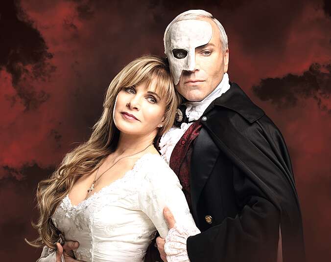 Ein grauhaariger Mann in schwarzem Umhang und mit einer weißen Maske über eine Gesichtshälfte hält eine blonde, langhaarige Frau in weißem Ballkleid von hinten im Arm.