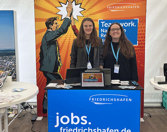 Zwei Frauen stehen hinter Counter vor Videowand mit Werbung für Jobs bei der Stadtverwaltung Friedrichshafen.