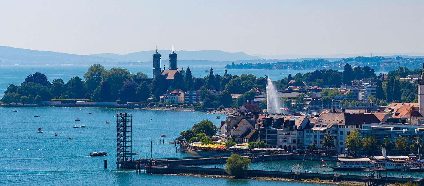 Panoramabild vom See aus auf die Uferpromenade mit Moleturm, Seefontäne, Schloss und im Hintergrund der Sicht auf Fischbach
