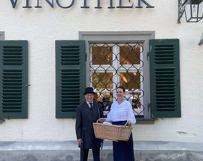 Frau mit Wäschekorb und Mann in Kleidung des 20. Jahrhunderts stehen vor der Vinothek Schloss Friedrichshafen.