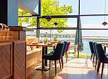 Modern eingerichtes Restaurant mit bunten Stühlen vor einer Glasfront mit Blick auf den Bodensee und ein Schiff.