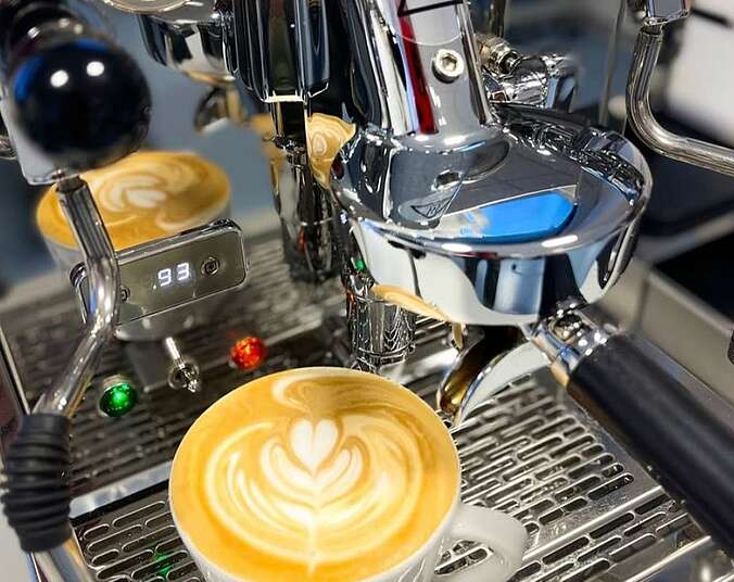 Eine glänzende Siebträger-Kaffeemaschine aus Edelstahl, mit sichtbaren Druck- und Temperaturanzeigern. Ein Cappuccino mit Herz aus Milchschaum steht auf der Maschine.