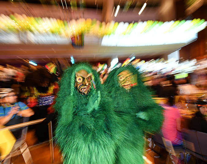 Zwei Seewaldkobolde mit Masken und grünem Häs zwischen Tischen, an denen Menschen sitzen