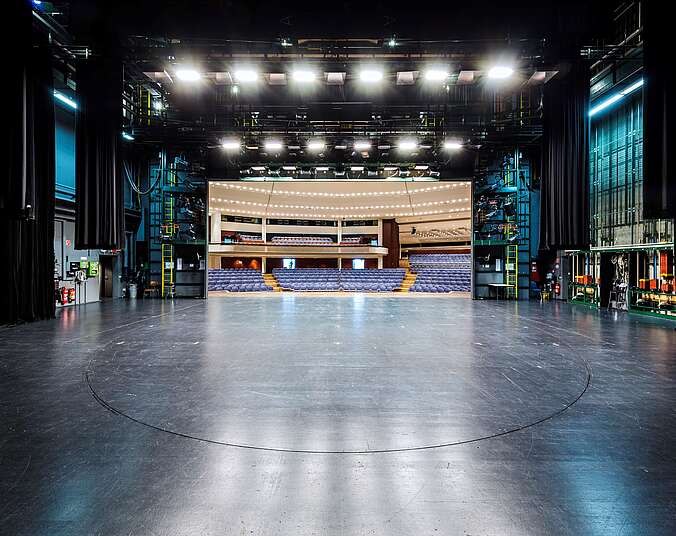 Blick von der Rückwand einer Bühne, über die leere Bühnenfläche in einen vollbestuhlten Saal mit Empore. An den Seitenwänden der Bühne sind Seilzüge, gelbe Leitern und Bühnentechnik zu sehen.