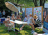 Jugendliche sitzen an Biergarnitur-Stand auf Wiese mit Plakatstellwänden, Rollup und Sonnenschirm