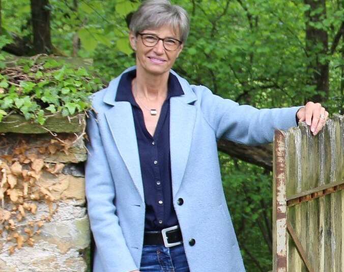 Eine Frau mit kurzen grauen Haaren, schwarzer Brille in dunkelblauem Hemd und hellblauem Kurzmantel lehnt mit ihrer rechten Schulter an einer mit Efeu bewachsenen Steinmauer, ihr linker Arm liegt locker auf einem Holzlattentor auf.