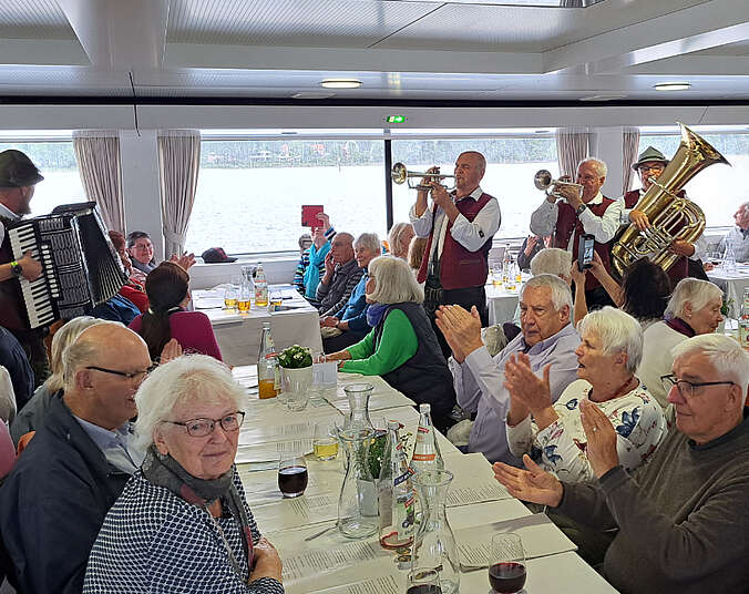 Männer und Frauen sitzen an Tischen und klatschen. Im Hintergrund spielen Musiker mit verschiedenen Instrumenten.
