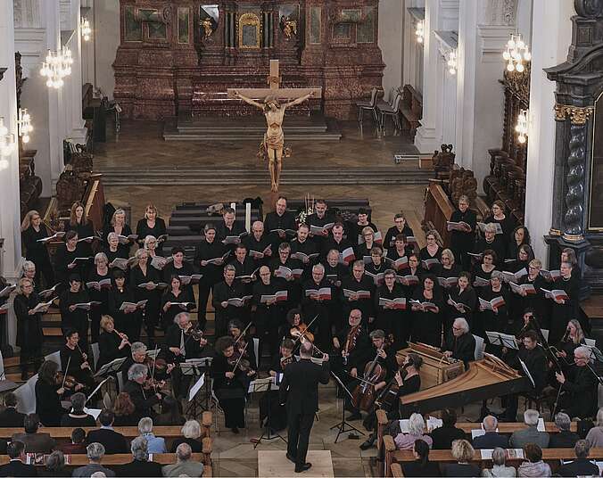 Von oben in den Altarraum einer Kirche fotografiert: Ein Chor im Halbkreis stehnd mit Notenmappen in den Händen, im Hintergrund ein goldenes Kreuz mit einem daranhängenden Jesus. Die vorderen Kirchenbänke sind mit Zuschauern besetzt.