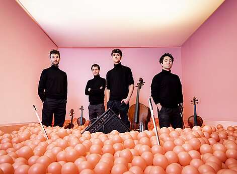Musiker des vision string quartet stehen mit ihren Instrumenten in einem rötlich beleuchteten Raum.