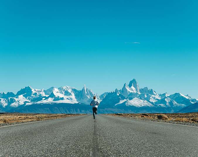 Ein Mann von hinten fotografiert läuft auf einer einsamen Landstraße durch eine karge Landschaft auf eine Gebirgskette zu.