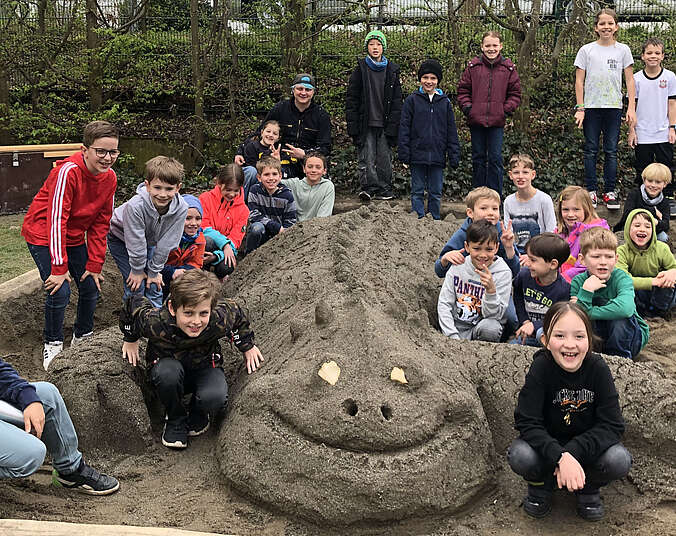 Lachende Kinder stehen um einen aus Sand gebauten Drache.