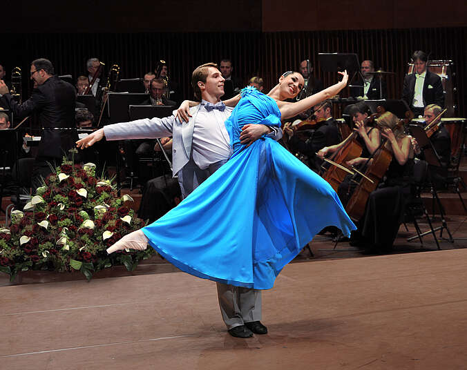 Ein junges Tanzpaar tanzt eine Hebefigur vor einem Orchester. Der Tänzer trägt einen hellgrauen Anzug, die Tänzerin ein hellblaues Ballkleid.